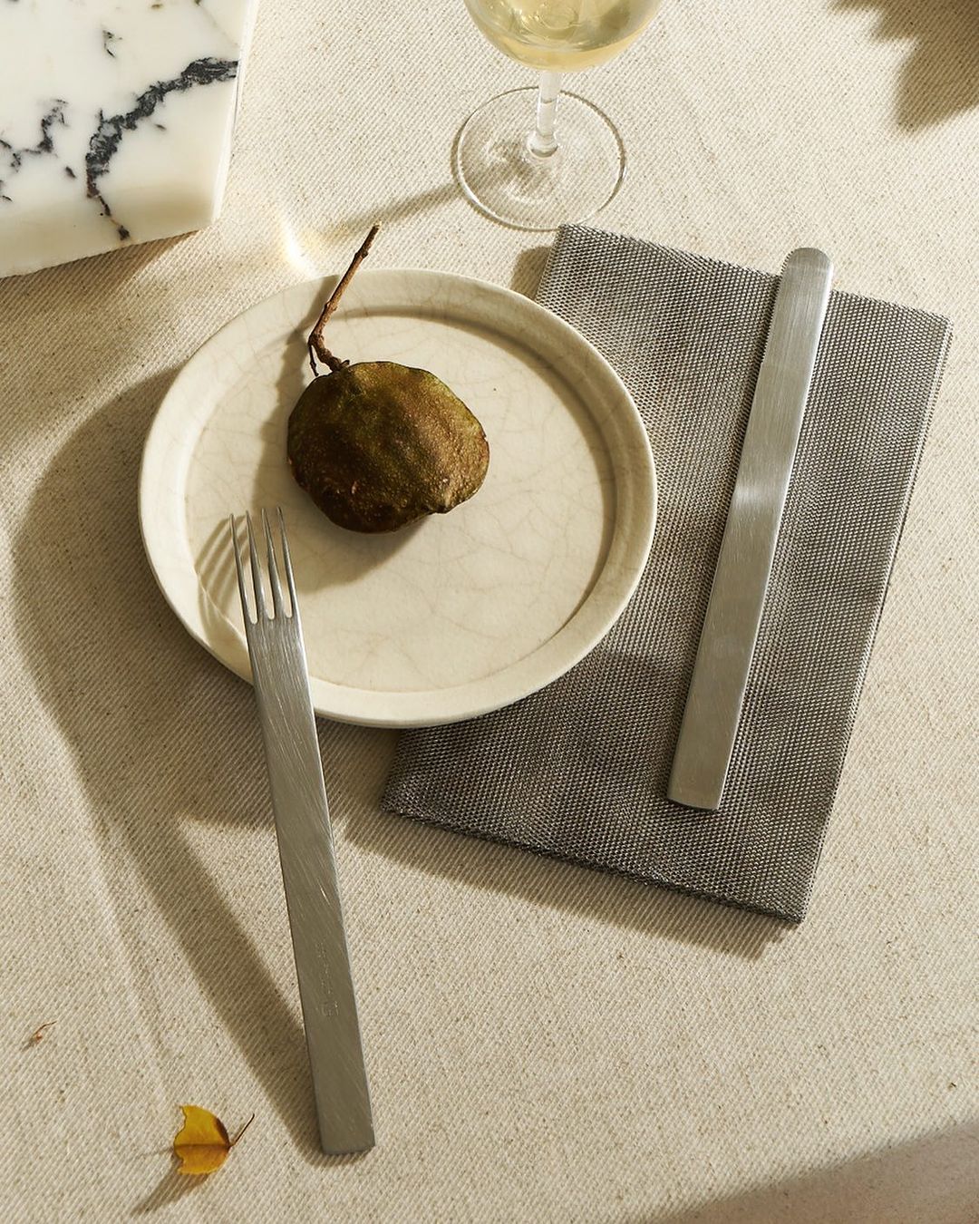 Still life featuring a pear on a plate with silverware, a napkin, and a glass on a linen tablecloth. Neutral tones and soft light.