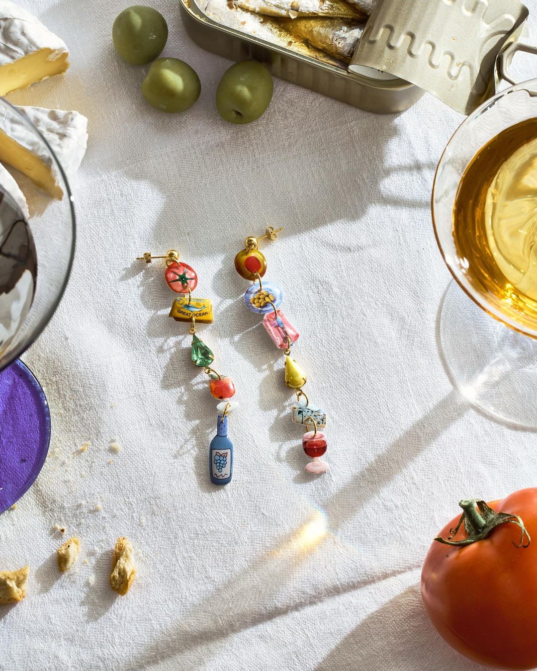 Eye-level shot of dangling food and drink earrings displayed with sardines, cheese, olives, a tomato, and drinks against white fabric.