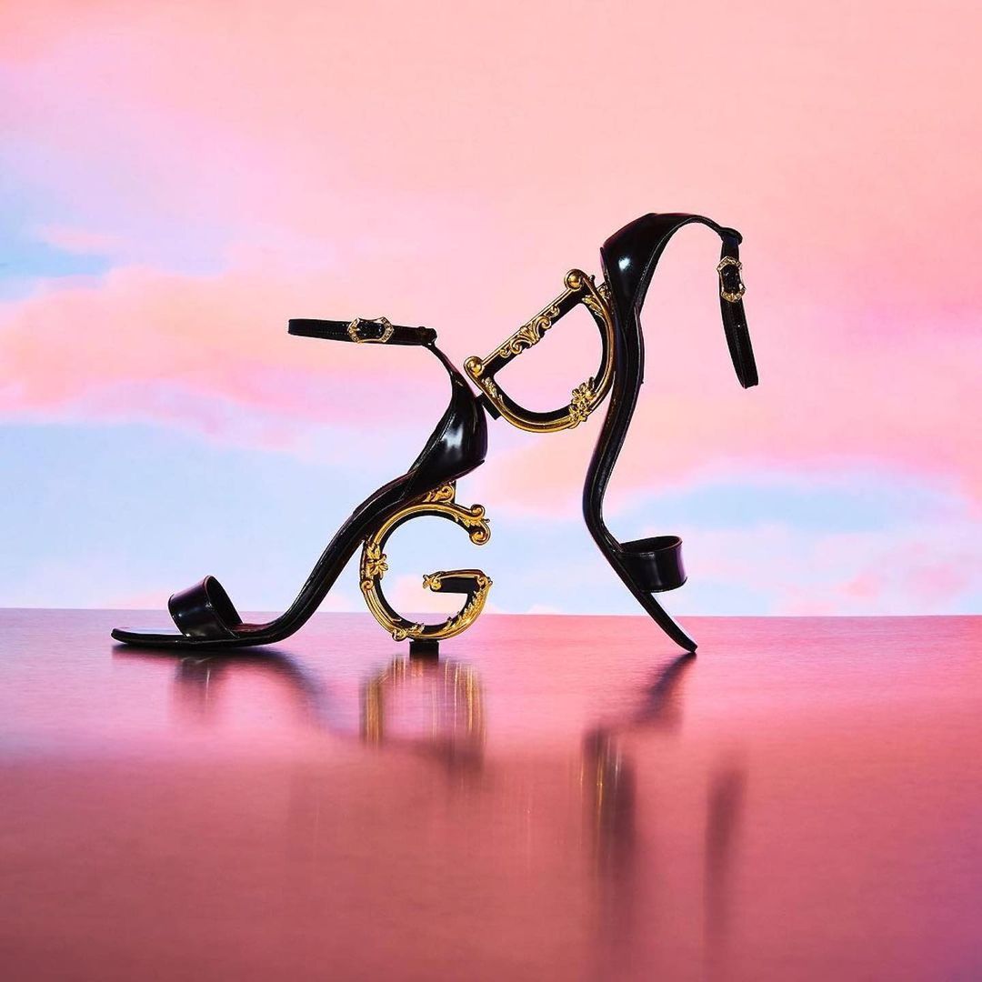 Black high-heeled sandal with gold embellishments on a reflective surface, set against a pastel gradient sky backdrop.