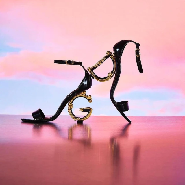 Black high-heeled sandal with gold embellishments on a reflective surface, set against a pastel gradient sky backdrop.