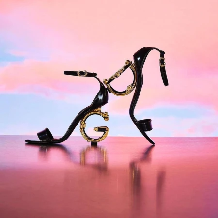 Black high-heeled sandal with gold embellishments on a reflective surface, set against a pastel gradient sky backdrop.