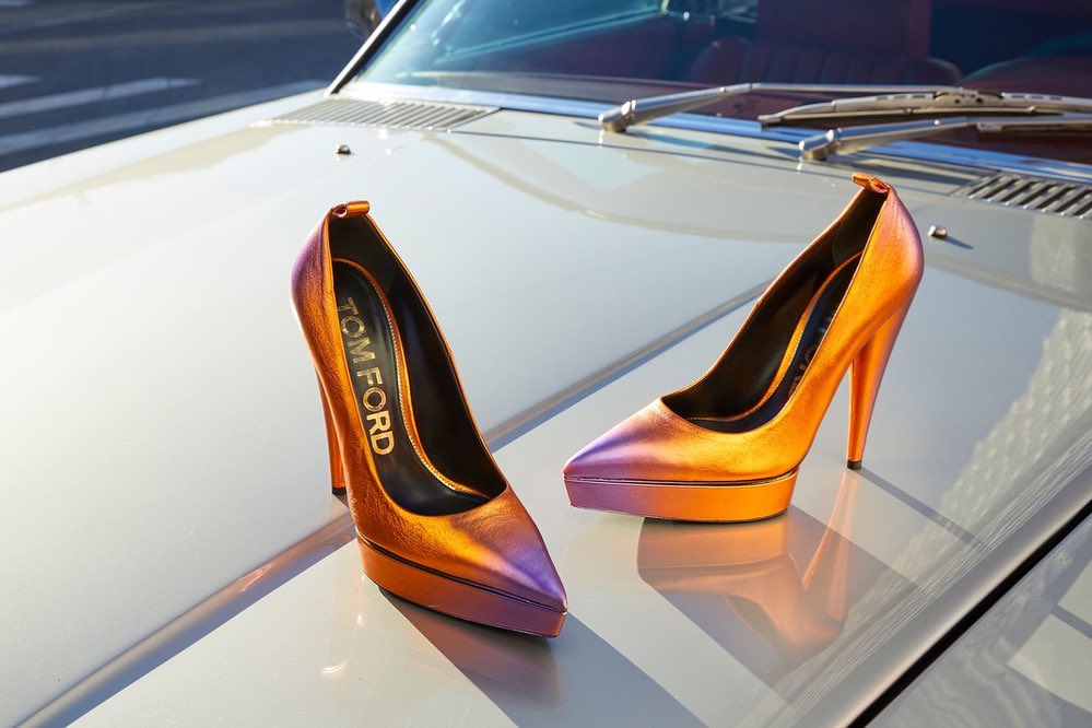 Two orange and purple Tom Ford stiletto high-heel shoes sit on the hood of a vintage car with chrome trim in a fashion shot.