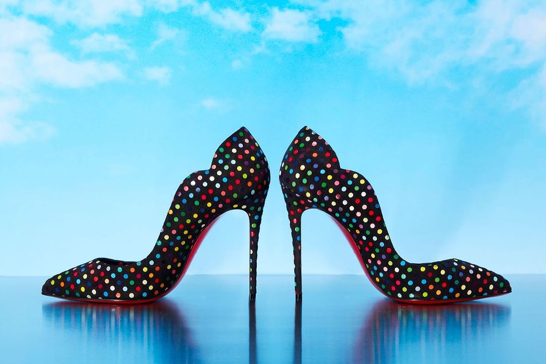 Pair of black high heel shoes covered in multi-colored polka dots with a red sole on a reflective surface with a blue sky background.