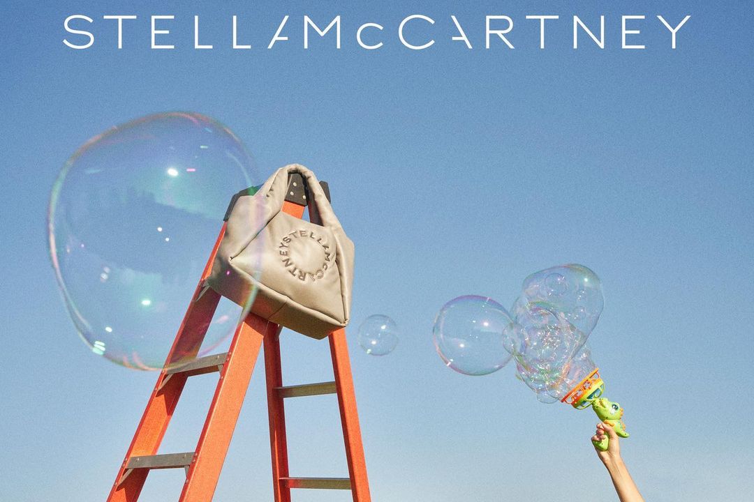 Stella McCartney bag on an orange ladder against a blue sky with soap bubbles blown from a toy bubble gun.  Outdoor setting.