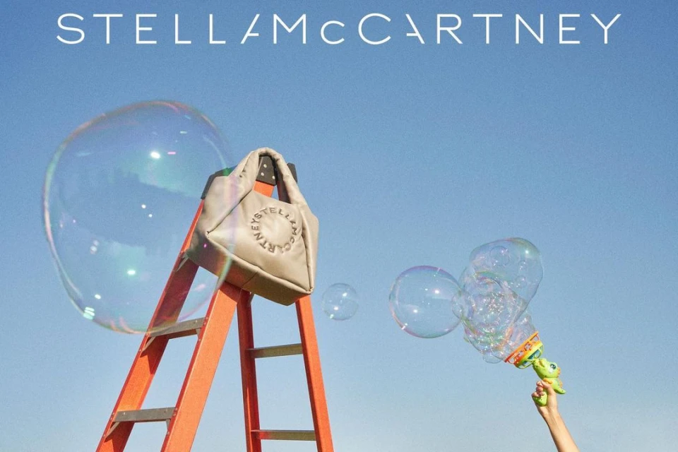 Stella McCartney bag on an orange ladder against a blue sky with soap bubbles blown from a toy bubble gun.  Outdoor setting.