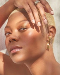 Close-up portrait of a Black woman with short blonde hair and gold eyeshadow, posing with her hand near her face.