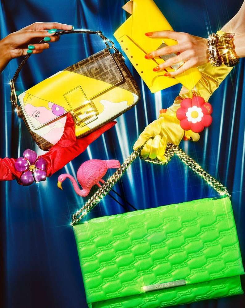Multiple designer handbags held by hands wearing colorful gloves and jewelry against a glossy blue background in a fashion shoot.