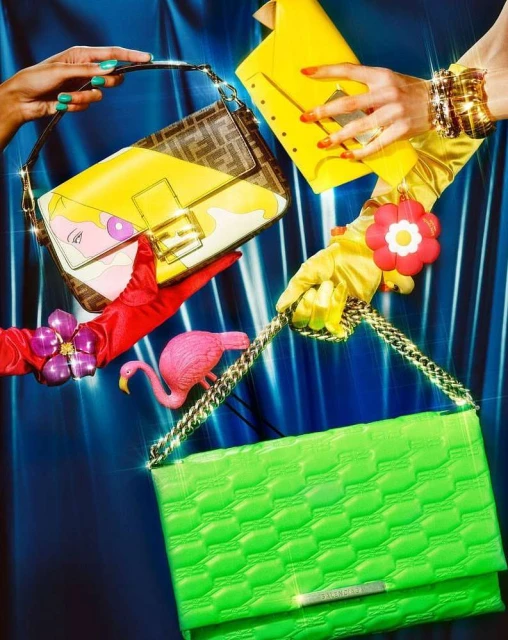 Multiple designer handbags held by hands wearing colorful gloves and jewelry against a glossy blue background in a fashion shoot.