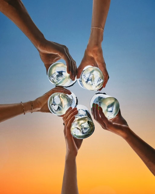 Cheers! Six arms raise clear glasses filled with ice against a bright blue and orange gradient sky in a celebratory toast, wrist jewelry visible.