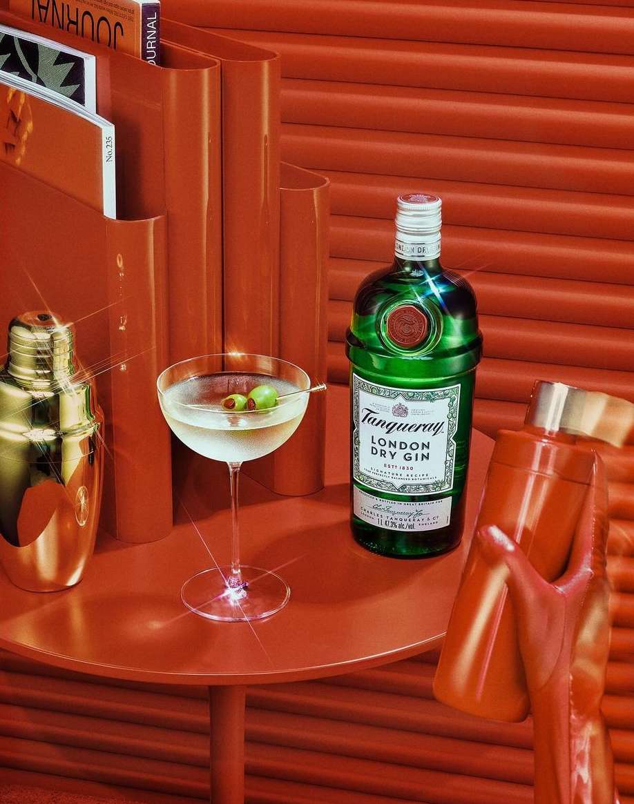 Green Tanqueray gin bottle, cocktail glass with olives, and gold cocktail shaker on an orange table with textured background.
