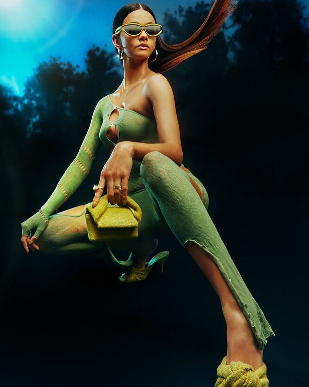 A woman in a one-shoulder green outfit poses with a green purse and matching heels against a cloudy blue and black backdrop.