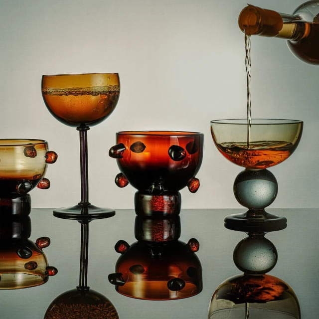 Wine being poured into an artful glass on a reflective surface, with three other similar glass designs in the background.