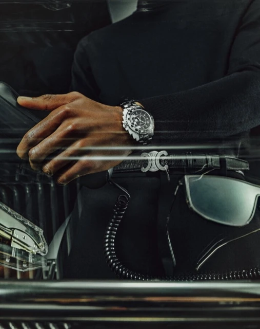 Close-up of a hand and wrist wearing a stainless-steel wristwatch, with a leather belt with a silver buckle and black fabric.