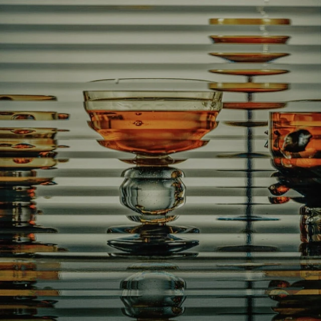 Close-up studio shot of two clear crystal glasses with amber liquid against a white venetian blind background and glass table.