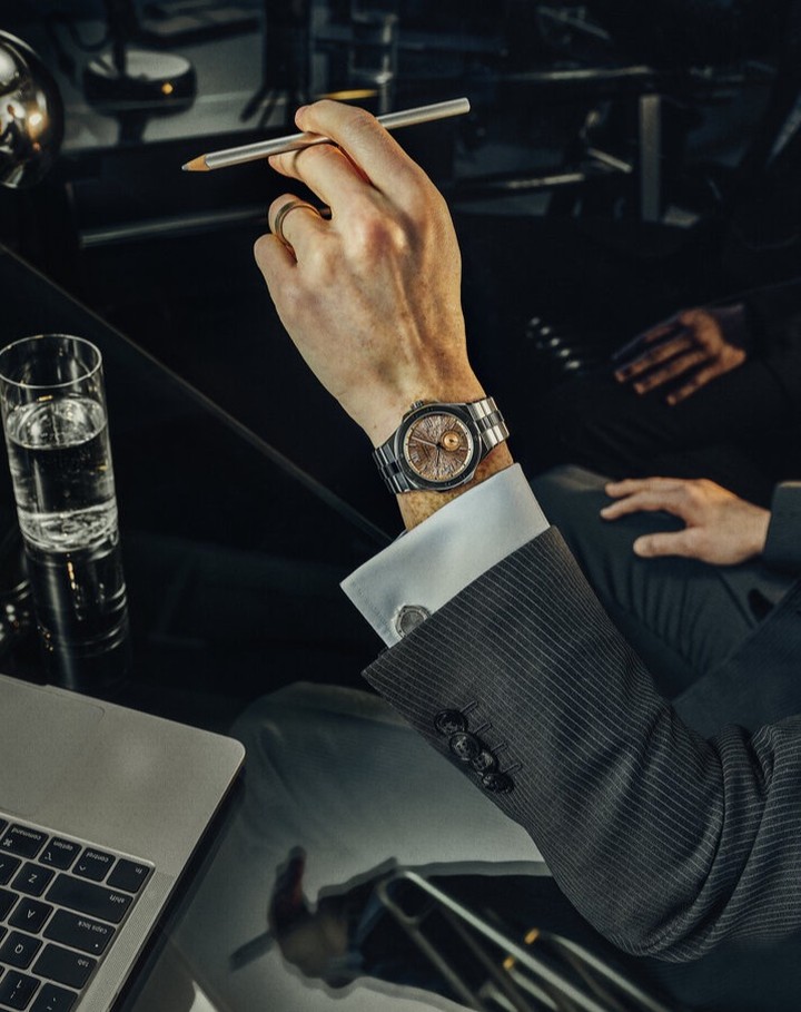Man in suit shows off wristwatch at desk with laptop, water glass, and pen. Focus on the watch and hand, in a dark, indoor setting.