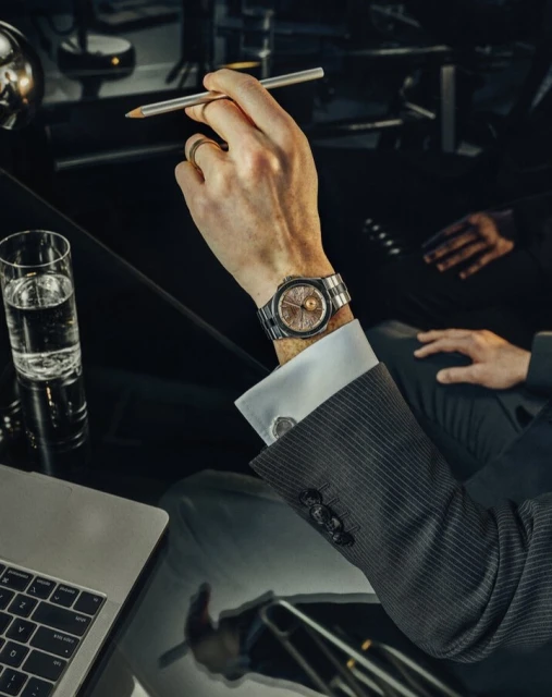 Man in suit shows off wristwatch at desk with laptop, water glass, and pen. Focus on the watch and hand, in a dark, indoor setting.