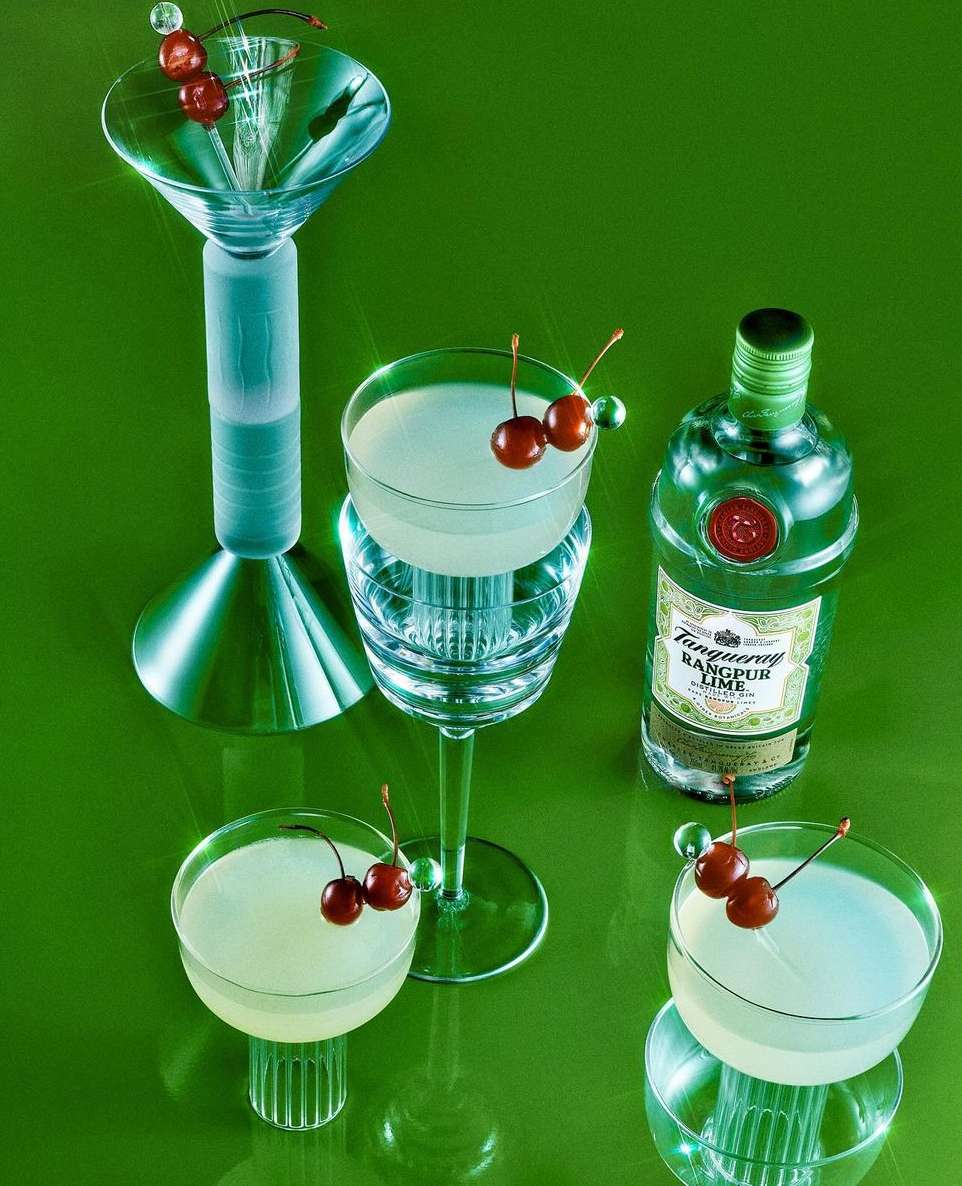 Still life of four cocktail glasses with cherries and a Tanqueray bottle on a green surface in a studio setting, product photography.