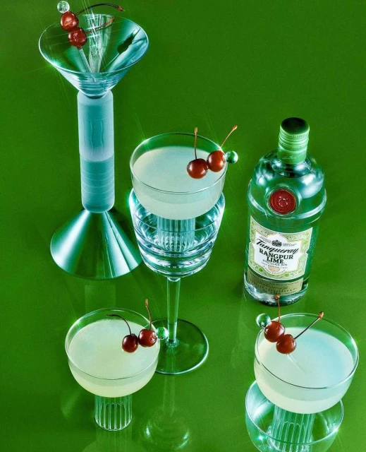 Still life of four cocktail glasses with cherries and a Tanqueray bottle on a green surface in a studio setting, product photography.