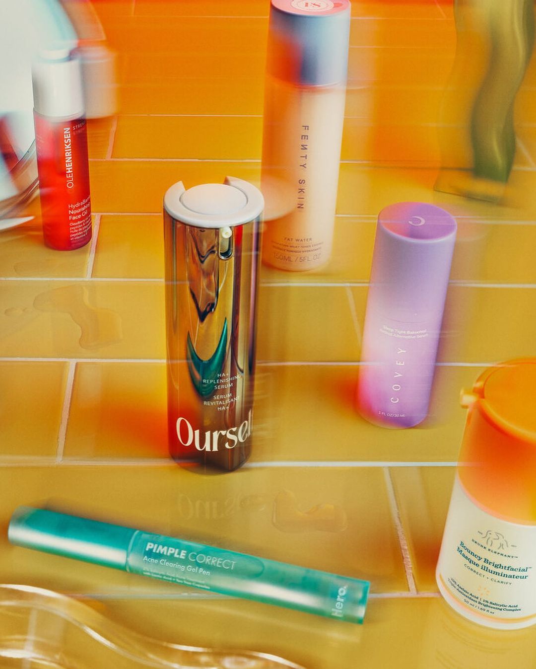 Several skincare products including serums and moisturizers from brands like Fenty Skin and Drunk Elephant on a yellow tiled surface.