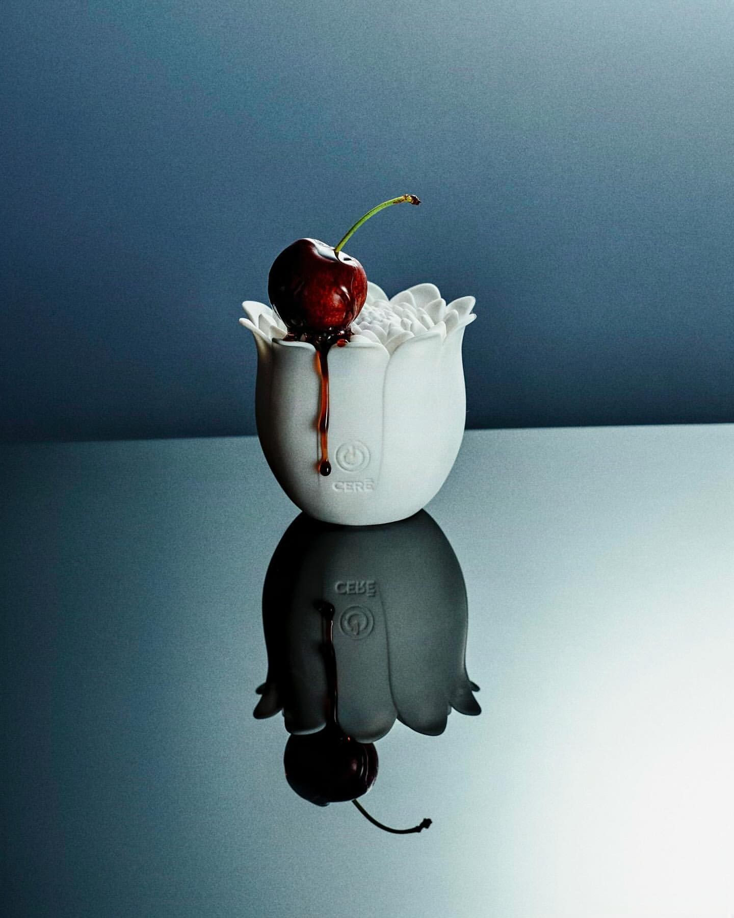 A dark red cherry sits atop a white, tulip-shaped container with a clear gel, set against a blue background with a visible reflection.