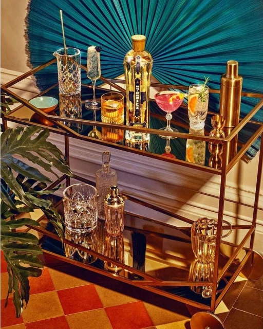 Stylish gold bar cart with St. Germain bottle, cocktail glasses, and bar tools against teal palm fan background, warm indoor lighting.
