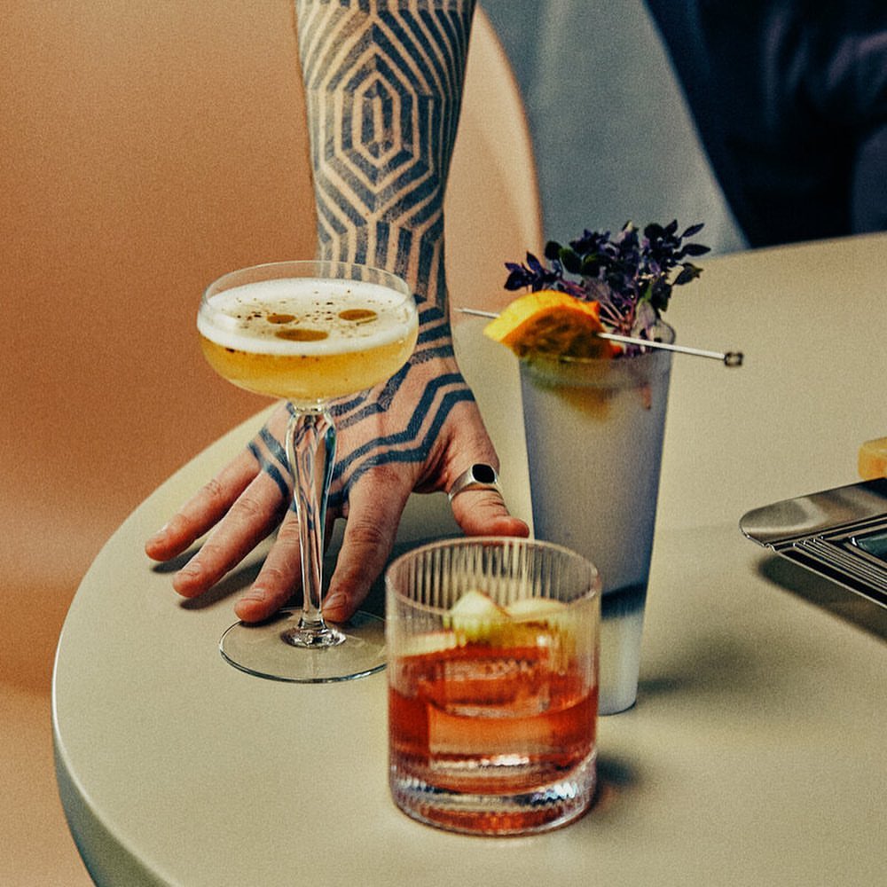 A close-up shot of three cocktails on a table, featuring a tattooed arm with geometric designs reaching for a stemmed glass.