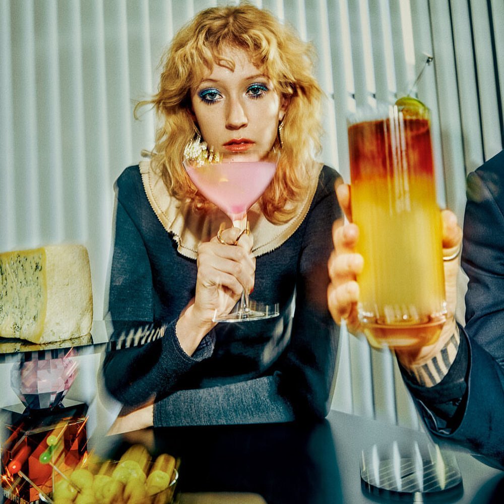 Woman holding a pink cocktail in front of white paneled wall and bar with an uncut piece of cheese and glass decor elements.