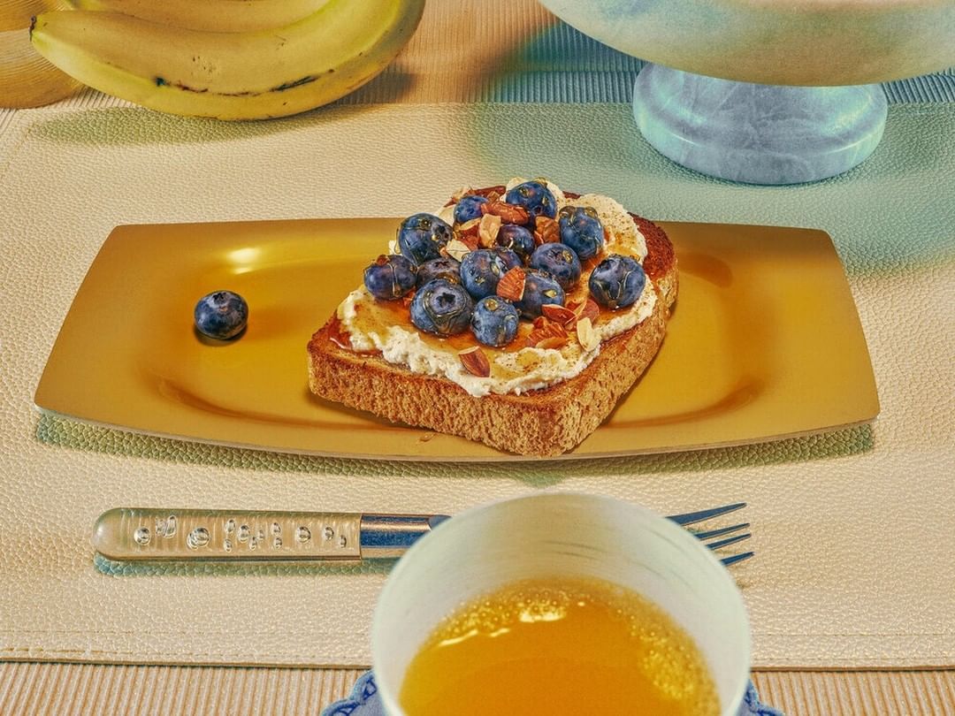 Close-up of a toast topped with cream cheese, blueberries, and sliced almonds served on a yellow plate with fruit and juice.