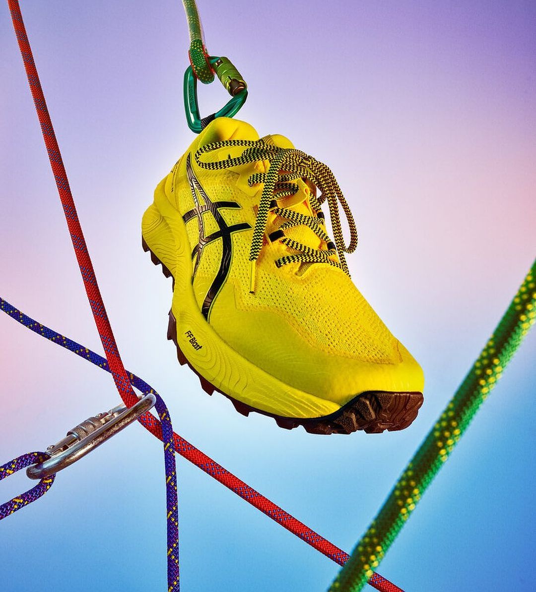 Bright yellow running shoe hangs suspended from a carabiner and colorful climbing ropes against a soft blue and purple gradient background.