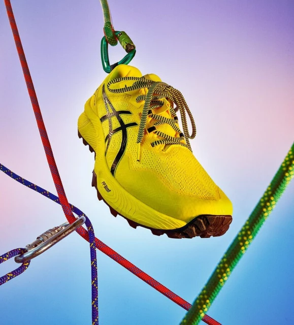 Bright yellow running shoe hangs suspended from a carabiner and colorful climbing ropes against a soft blue and purple gradient background.