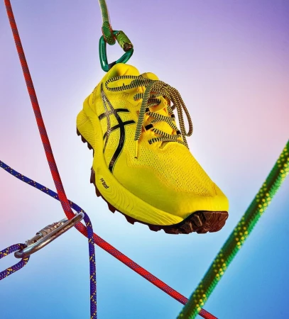 Bright yellow running shoe hangs suspended from a carabiner and colorful climbing ropes against a soft blue and purple gradient background.