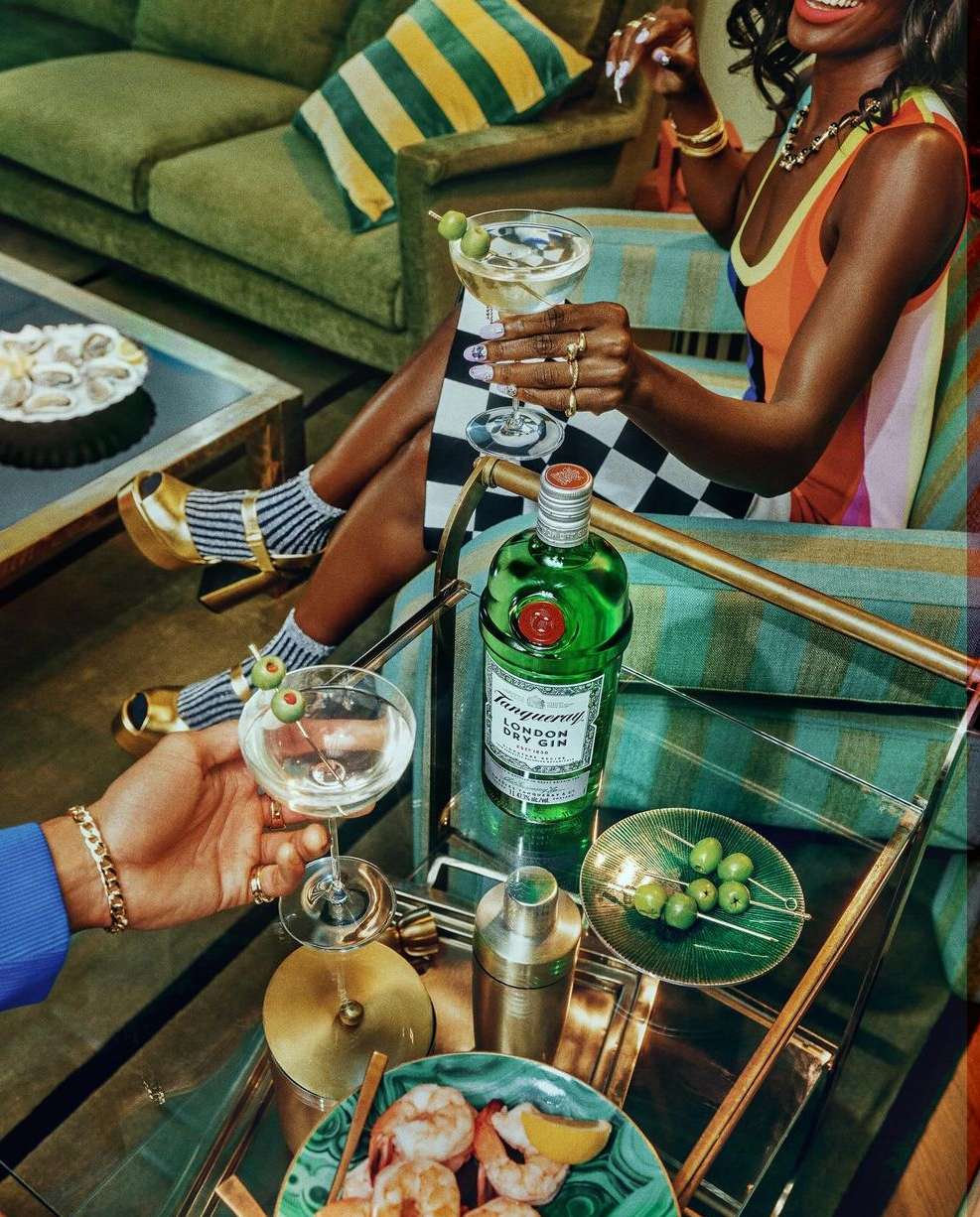 Woman in colorful dress holding a martini, cocktail on a bar cart with gin bottle, olives, shrimp, and a second hand holding a martini.