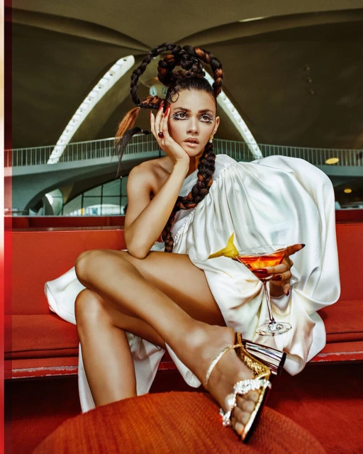 Elegant woman in architectural space, styled with ornate hair and wearing draped white fabric, holding a cocktail in a stemmed glass.