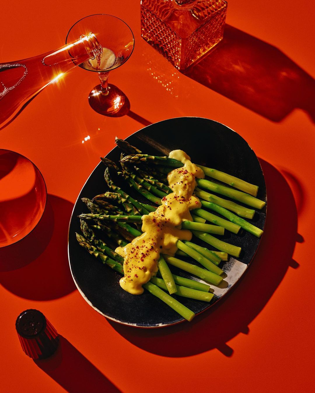 Overhead shot of asparagus with hollandaise sauce on black plate, wine bottle pouring into glass, on orange surface, studio lighting.