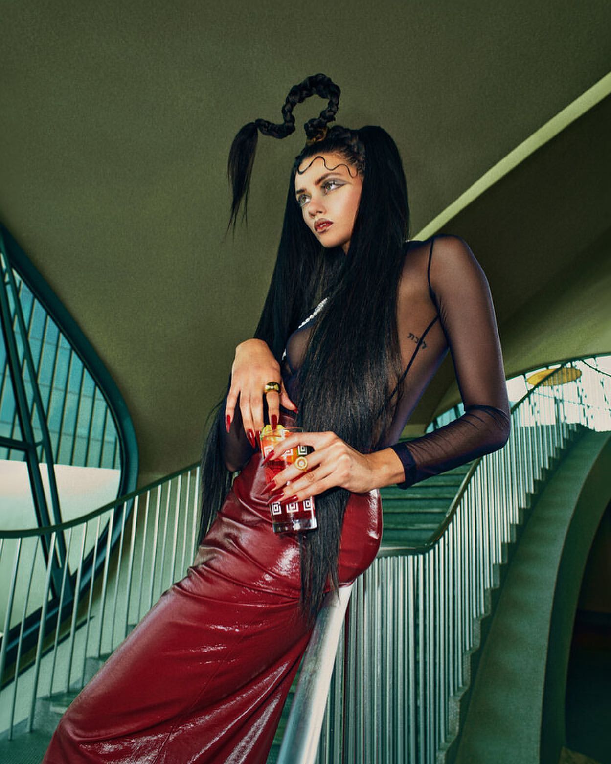 A young adult woman with intricately braided hair and a long red skirt leans against a staircase holding a glass of red beverage.