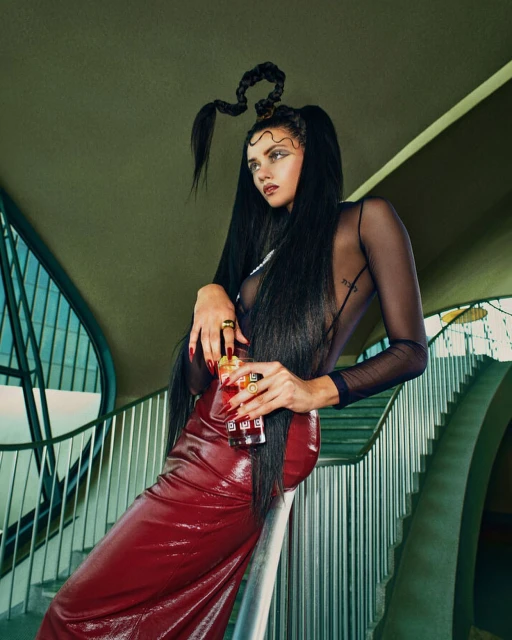 A young adult woman with intricately braided hair and a long red skirt leans against a staircase holding a glass of red beverage.
