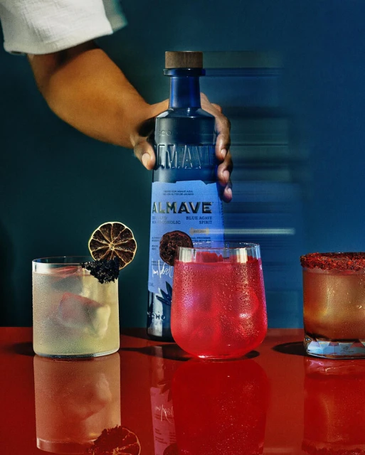 Cocktails and a bottle of Almave tequila are arranged on a red reflective surface against a teal backdrop with an arm reaching in.