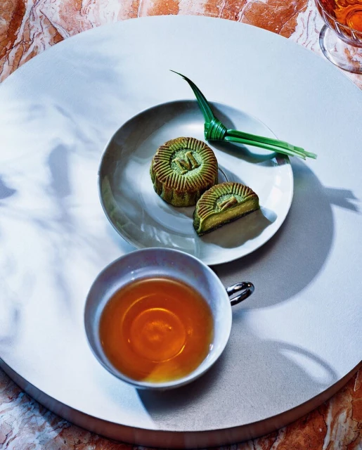 Overhead shot of two green-toned mooncakes on a small plate, along with a tea cup filled with amber liquid on a round surface.