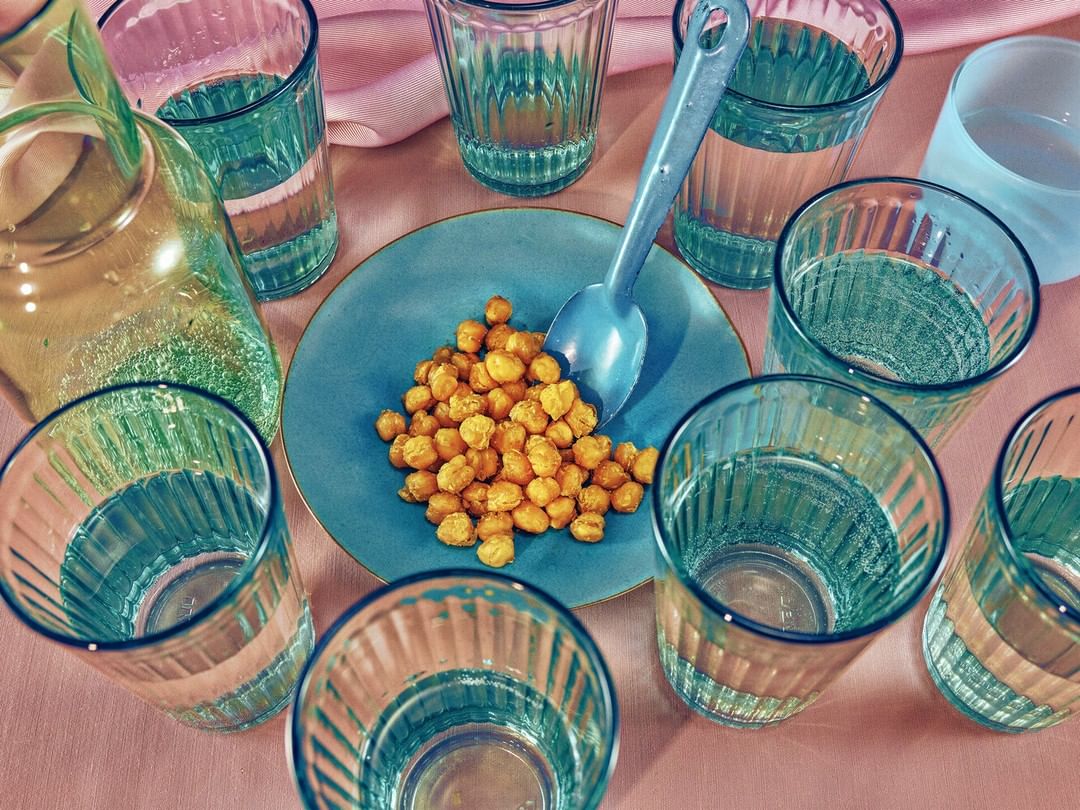 Overhead shot of blue rimmed glasses with water, a blue plate of chickpeas, and a blue spoon against a pink fabric surface.