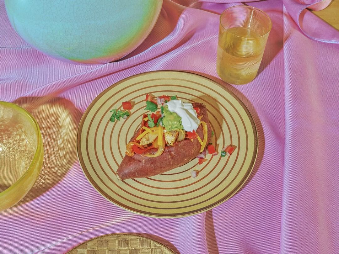 Overhead shot of a loaded sweet potato on a decorative plate with a glass of liquid, sitting on a pink satin cloth background.