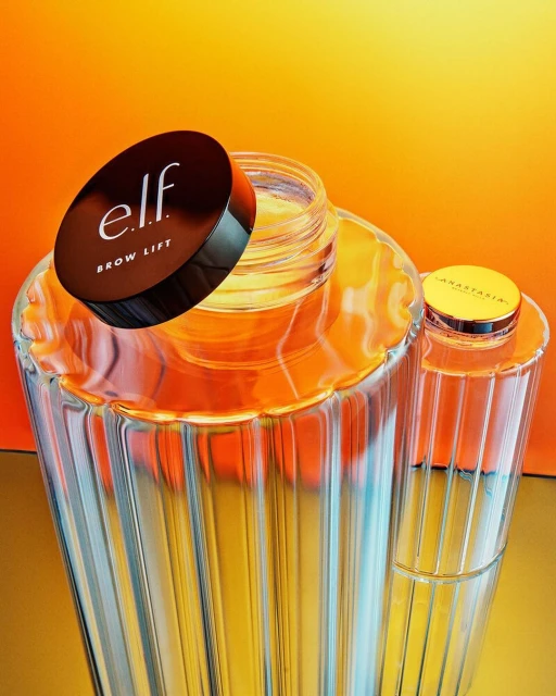 E.l.f. brow lift product next to an Anastasia Beverly Hills product, displayed in decorative glass on an orange to yellow gradient background.
