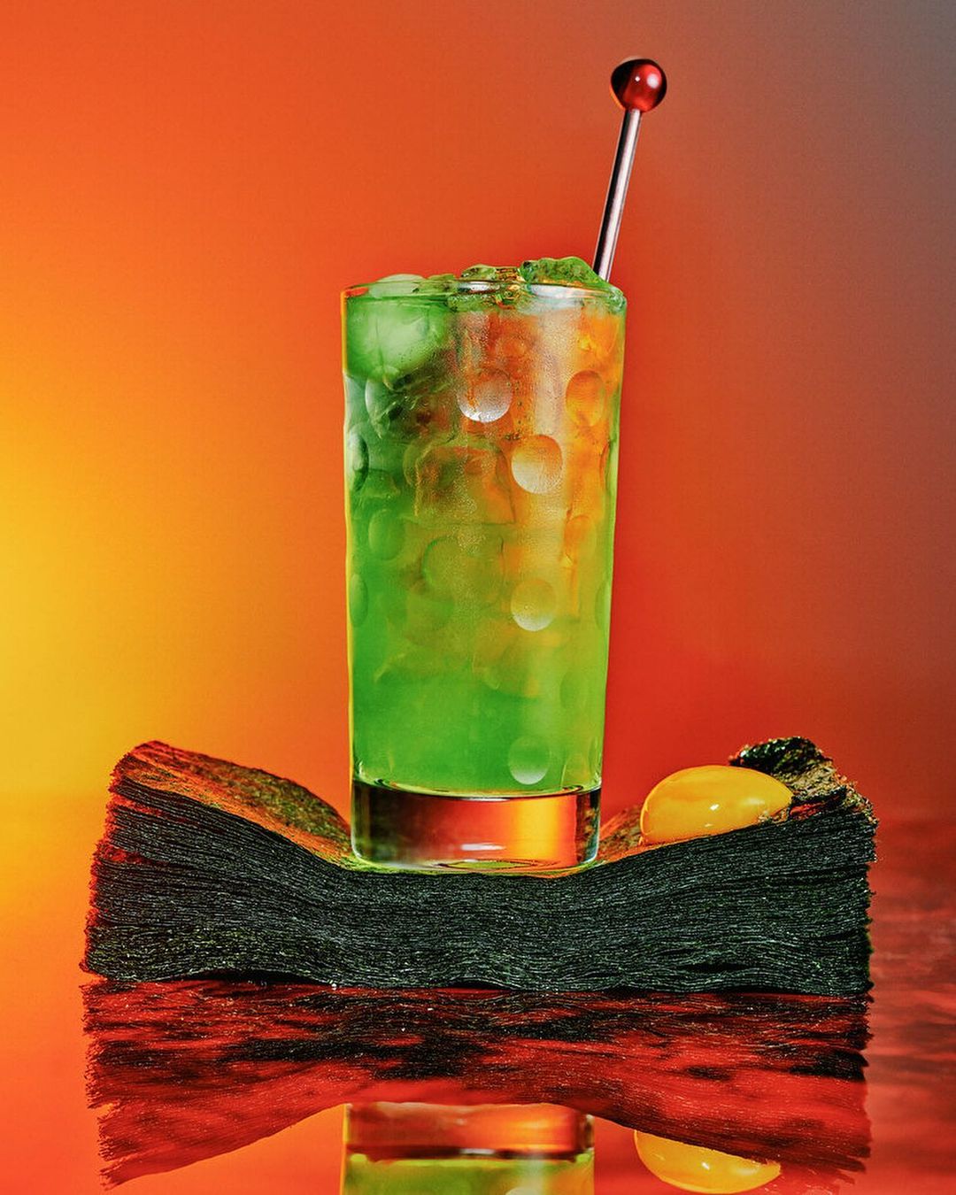 A green cocktail with ice is staged on a stack of black napkins with an orange background. The drink has a silver garnish.