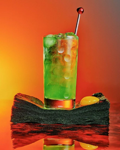 A green cocktail with ice is staged on a stack of black napkins with an orange background. The drink has a silver garnish.