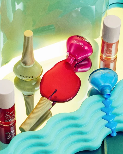 Gucci nail polish bottles in blue, pink, and green hues with spilled polish, and a turquoise textured wavy prop on a yellow surface.