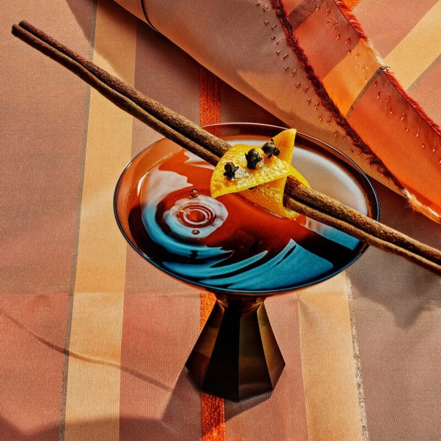 Elegant cocktail in a coupe glass with orange peel and cinnamon sticks. The drink is on a striped table cover.