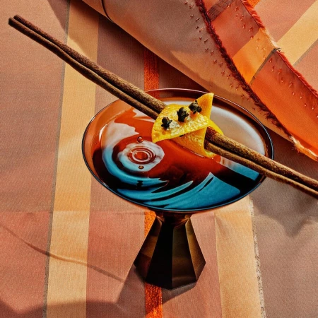 Elegant cocktail in a coupe glass with orange peel and cinnamon sticks. The drink is on a striped table cover.