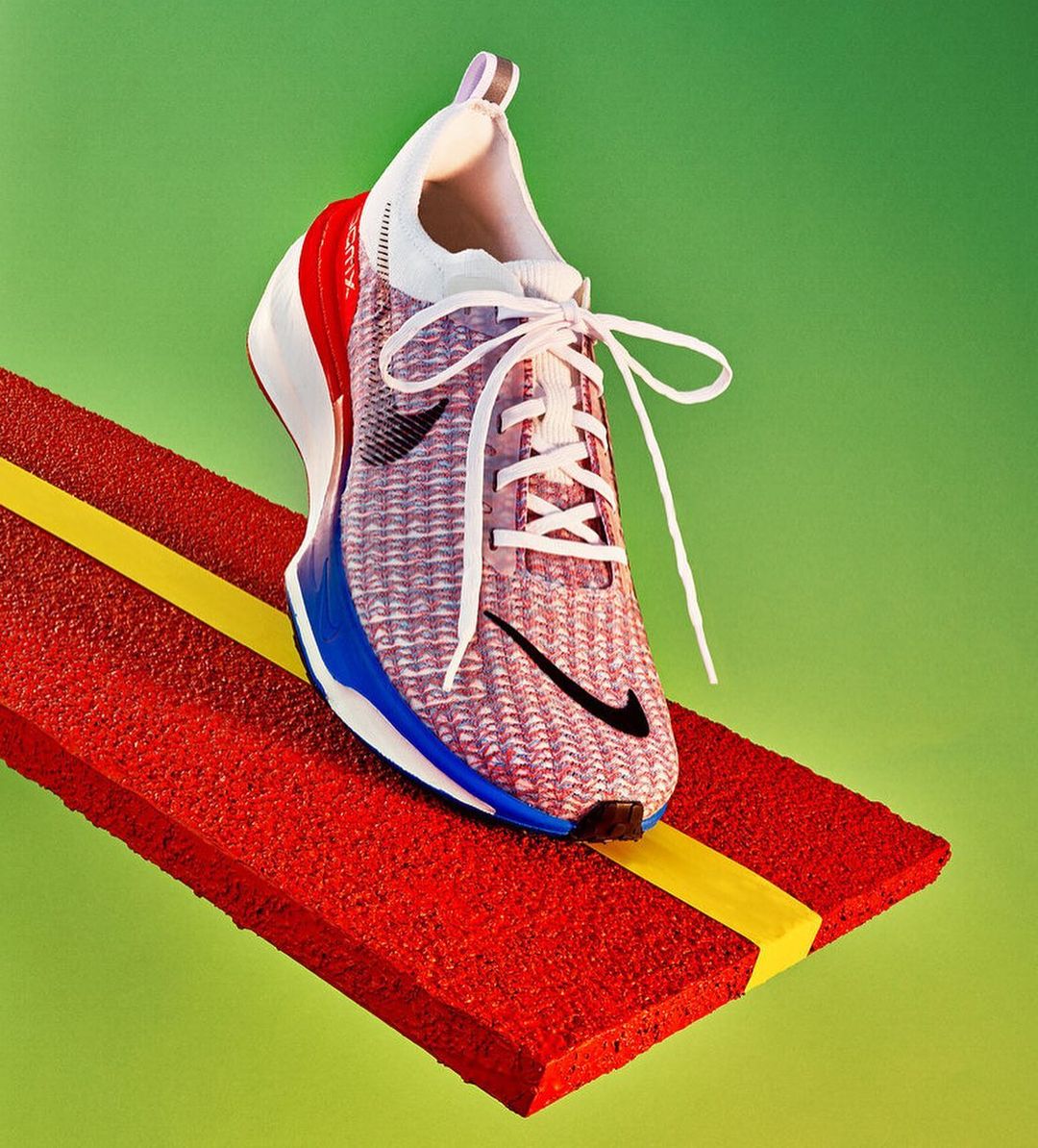 Close-up of a red, white, and blue Nike running shoe with white laces on a textured red platform against a bright green background.