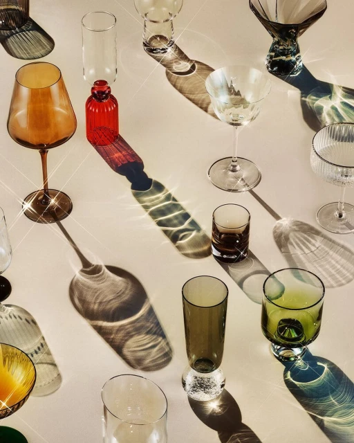 Still life shot of various coloured glassware including red, brown, green, and clear arranged on a light surface with shadows.