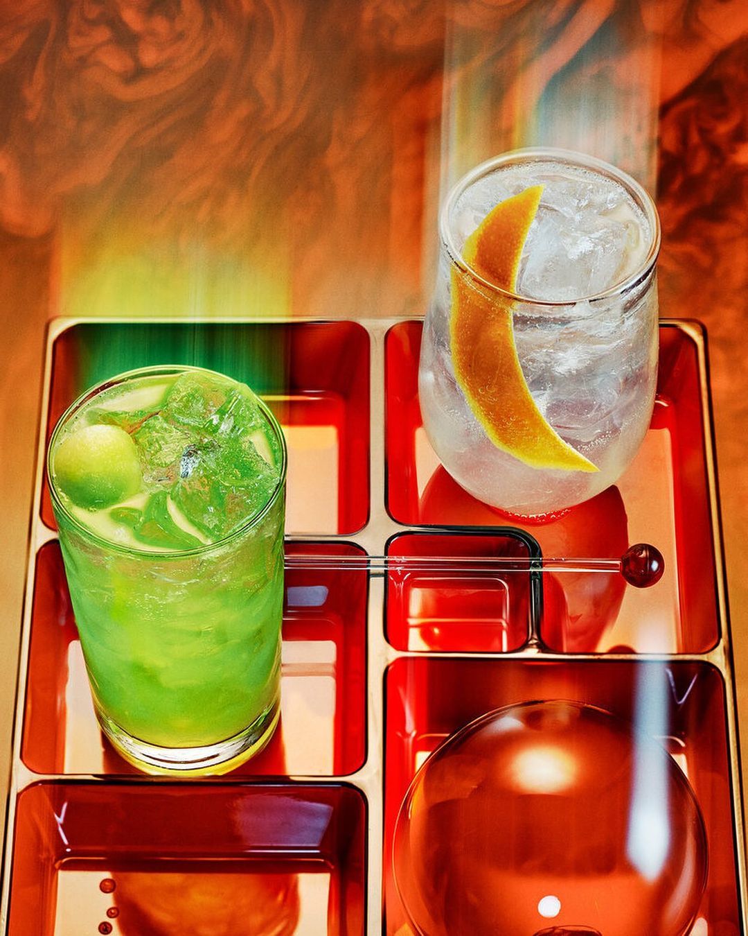 Two cocktails with ice cubes and orange peels are displayed on a divided tray with an orange and yellow smoke-like background.