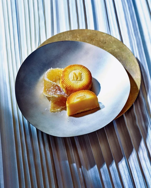 Elegant food still life featuring cut mooncakes with an 'M' logo, alongside candied fruit, served on silver and gold plates.